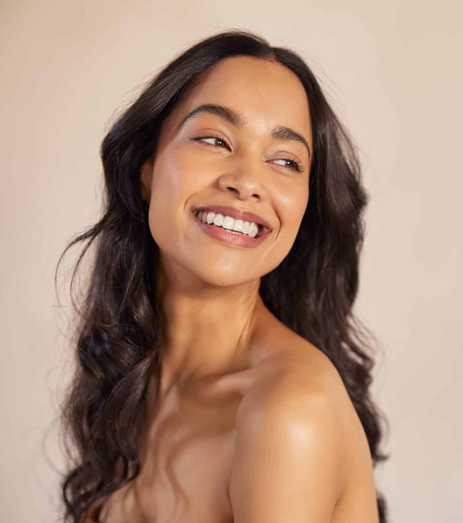 Smiling woman with long dark wavy hair and smooth, bare shoulders looks slightly away