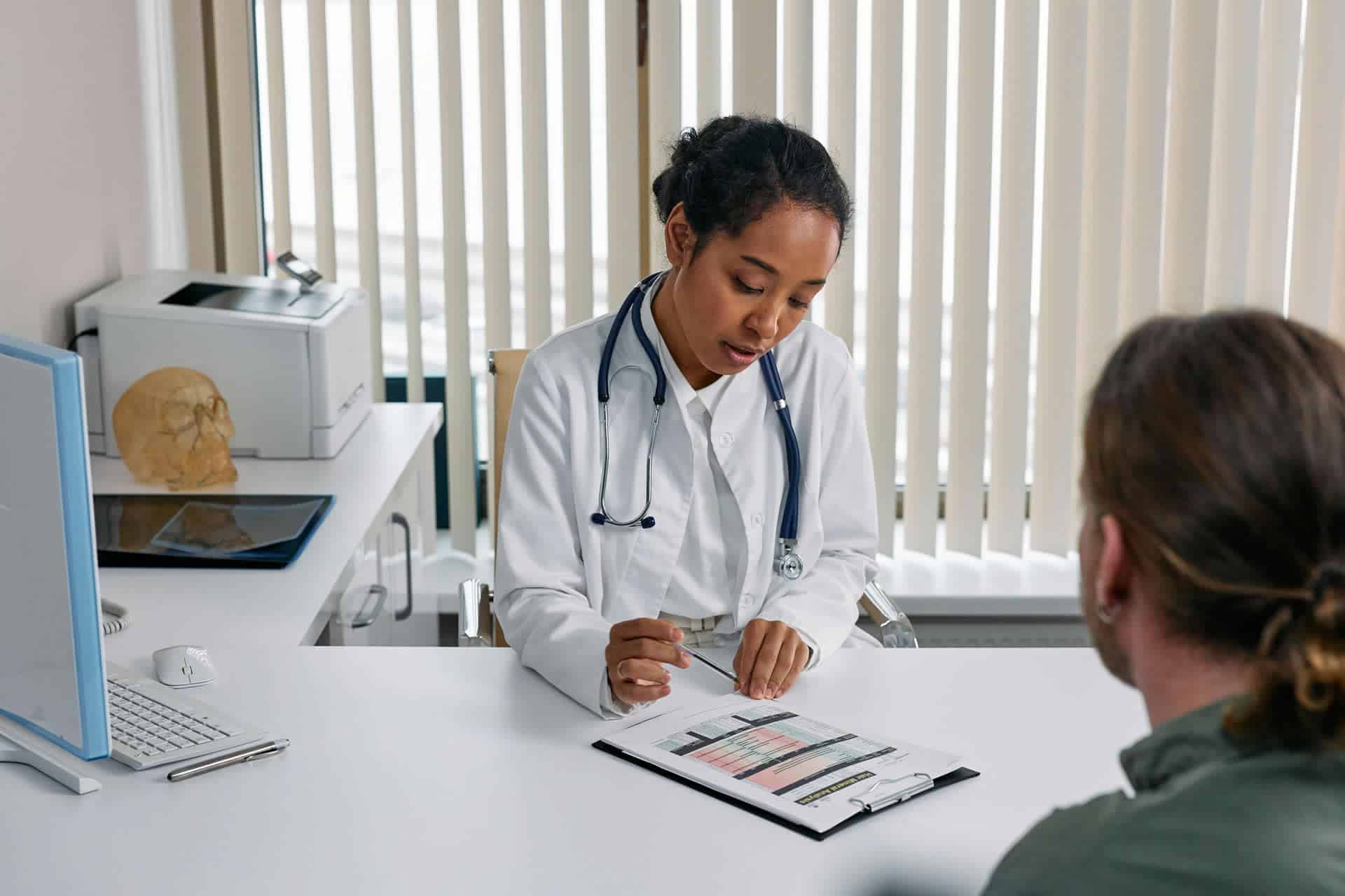 An African American female doctor in a white coat and stethoscope consults with a patient, pointing at a document on a clipboard