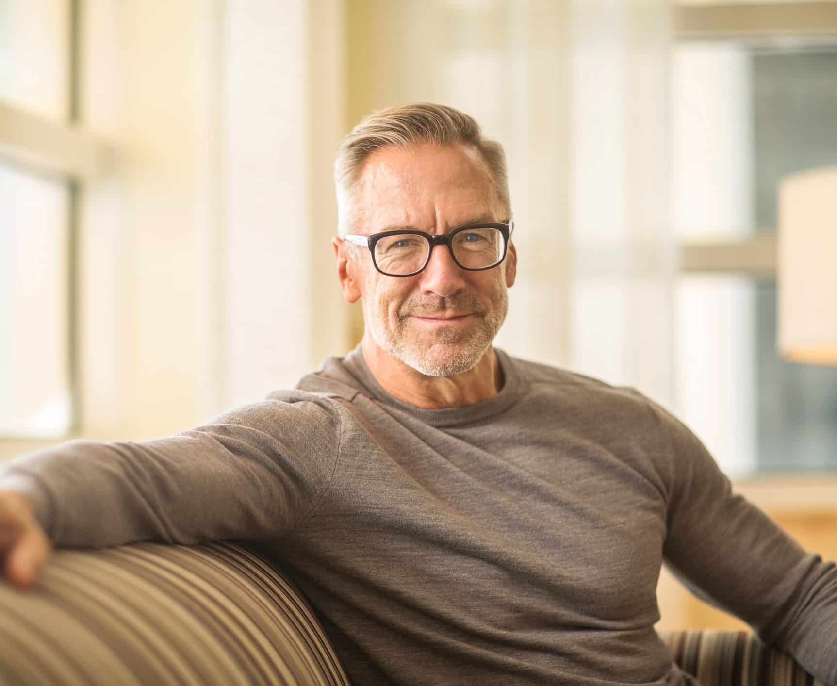A smiling mature man with grey hair and glasses sits comfortably on a striped couch, looking approachable