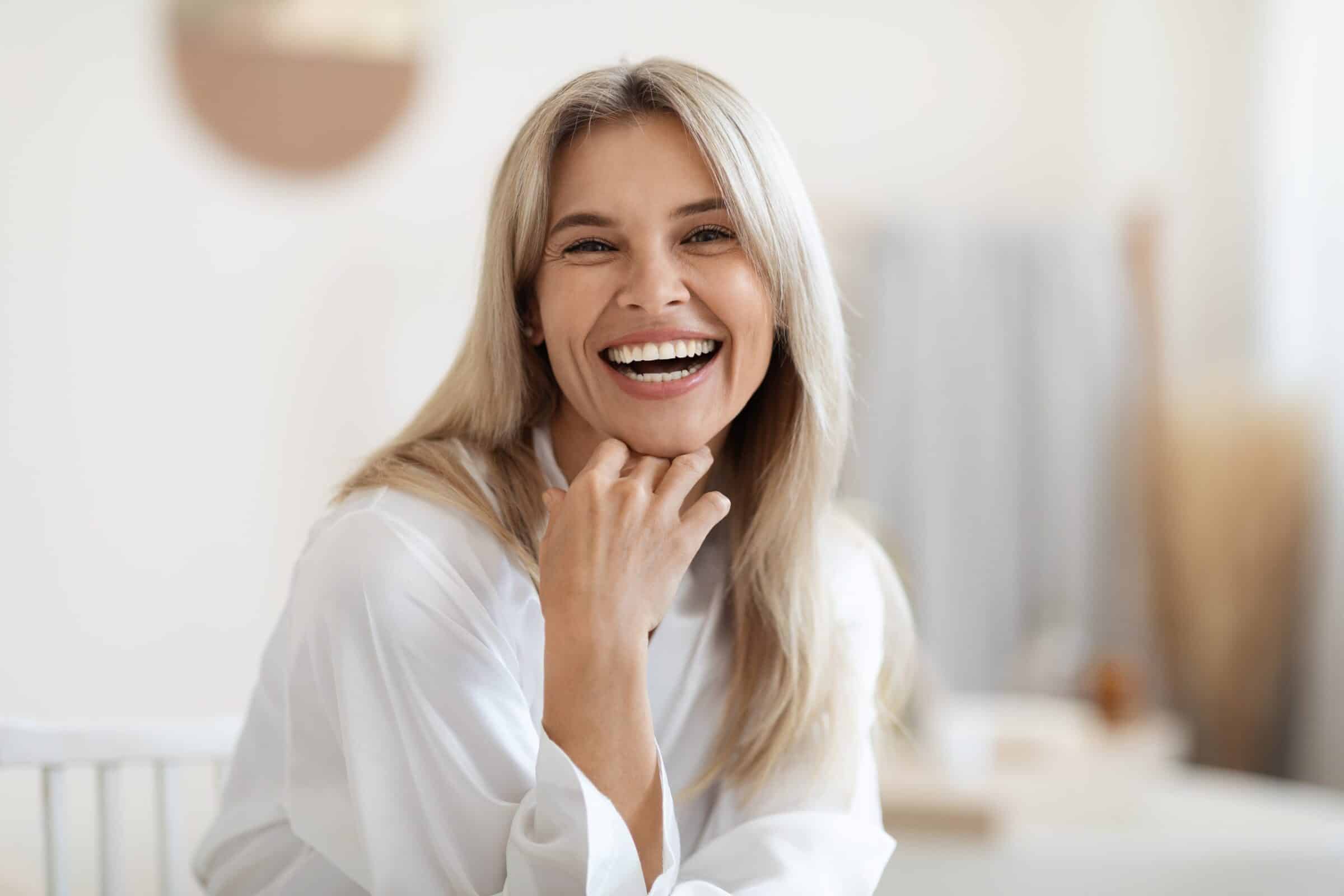 A happy blonde woman with a wide smile and bright teeth, laughing while resting her hand on her chin