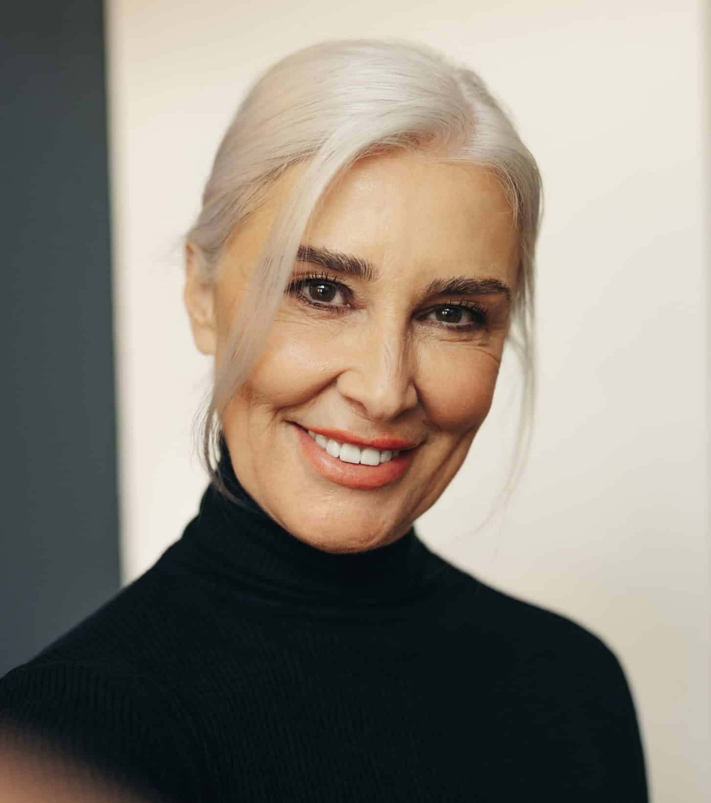 A smiling mature woman with elegant gray hair pulled back, wearing a black turtleneck, looks directly at the camera