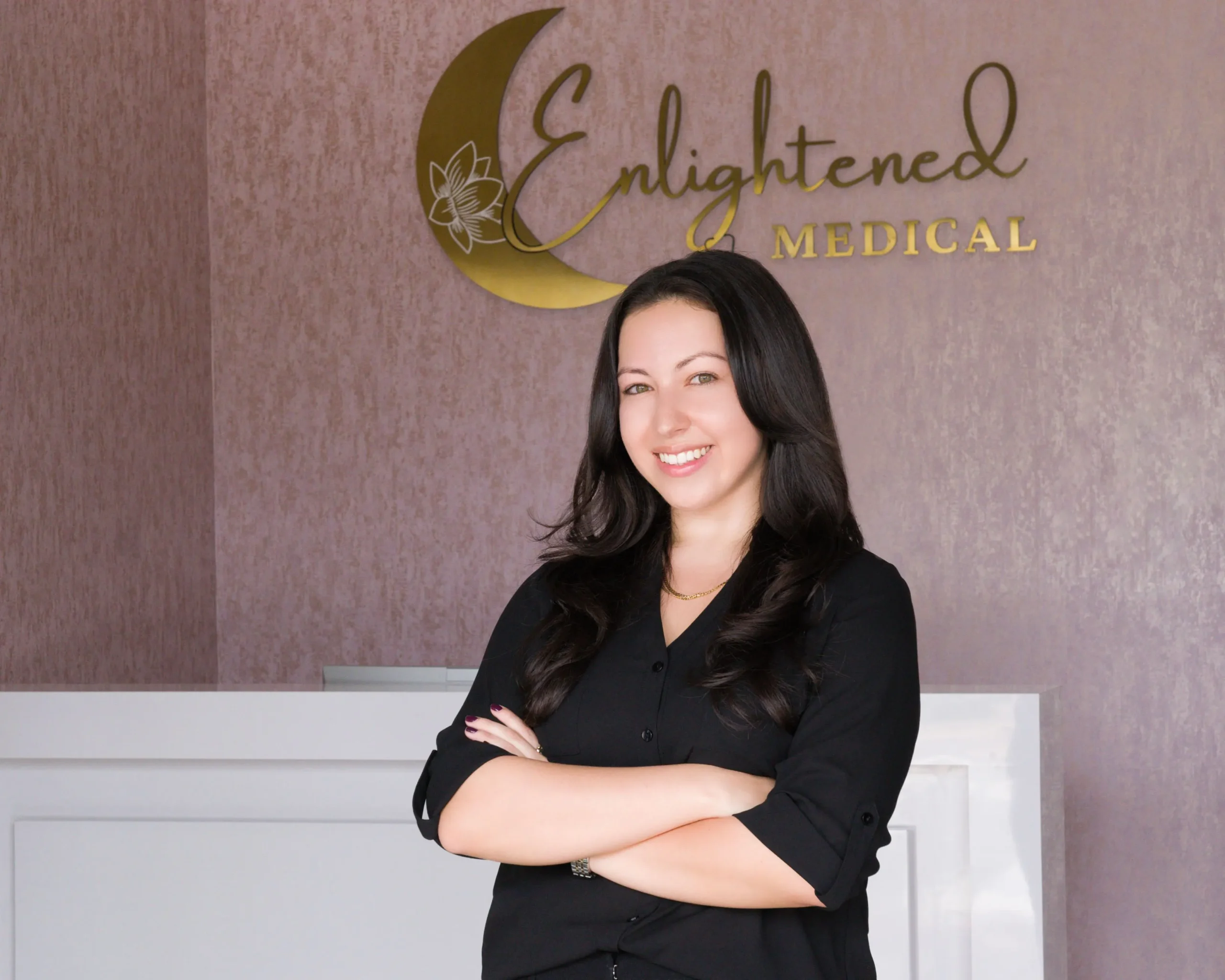 A smiling woman in a black shirt stands in front of a wall with the gold "Enlightened MEDICAL" logo