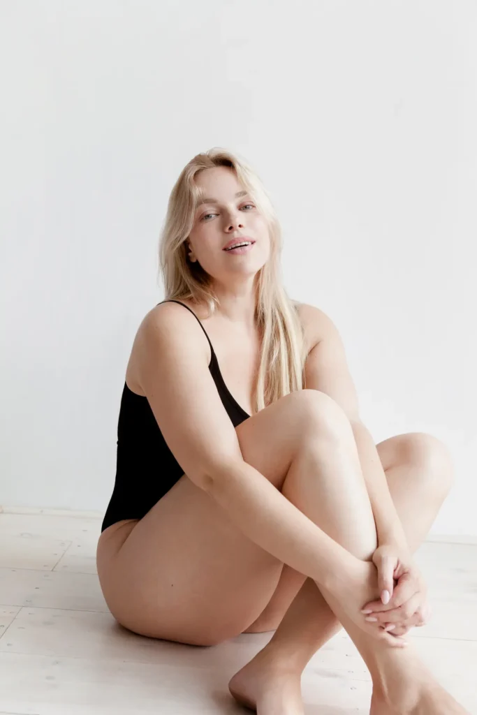 A blonde woman in a black one-piece swimsuit sits on a light wooden floor, looking at the camera. Her knees are bent, and she cradles her legs with her arms
