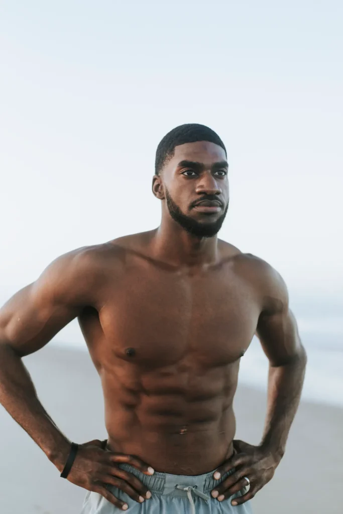 A shirtless Black man with a beard and muscular physique stands outdoors with hands on his hips