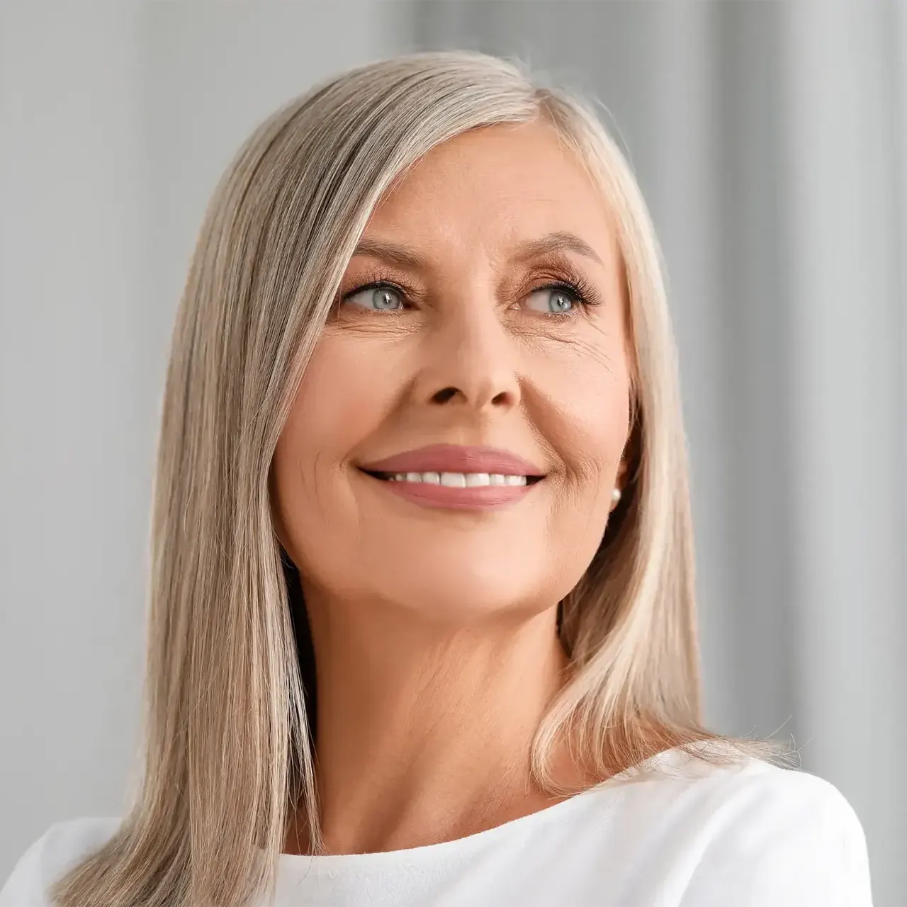 A smiling mature woman with long silver hair and blue eyes looks to the side, wearing a white top and a pearl earring