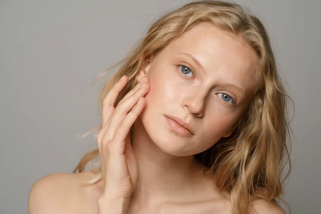 Young woman with blonde hair and blue eyes gently touches her cheek, emphasizing healthy, clear skin