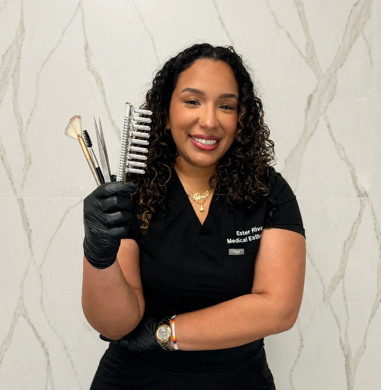 Ester Rivas Medical Esthetician wears black scrubs with FIGS tag, smiling and holding lash brushes, tweezers, and a comb