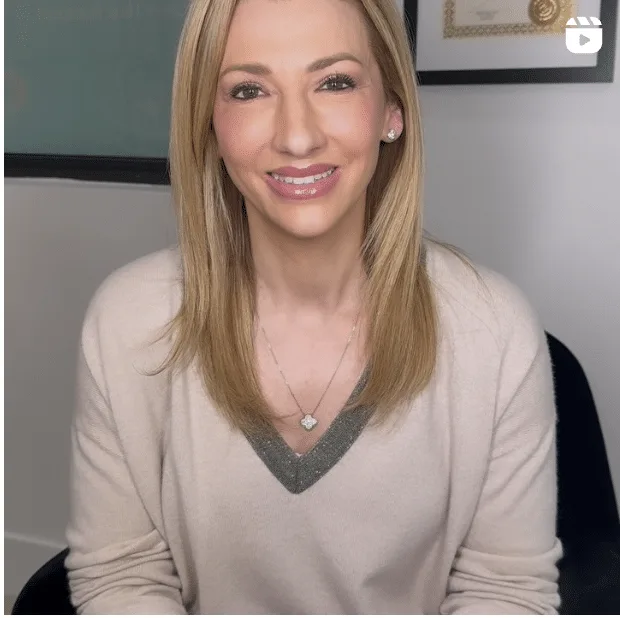 A smiling blonde woman wears a beige V-neck sweater, a clover pendant necklace, and diamond stud earrings