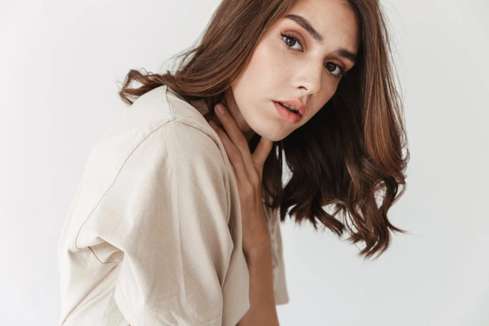 A young woman with brown wavy hair and a light-colored top looks at the camera, her hand gently touching her neck