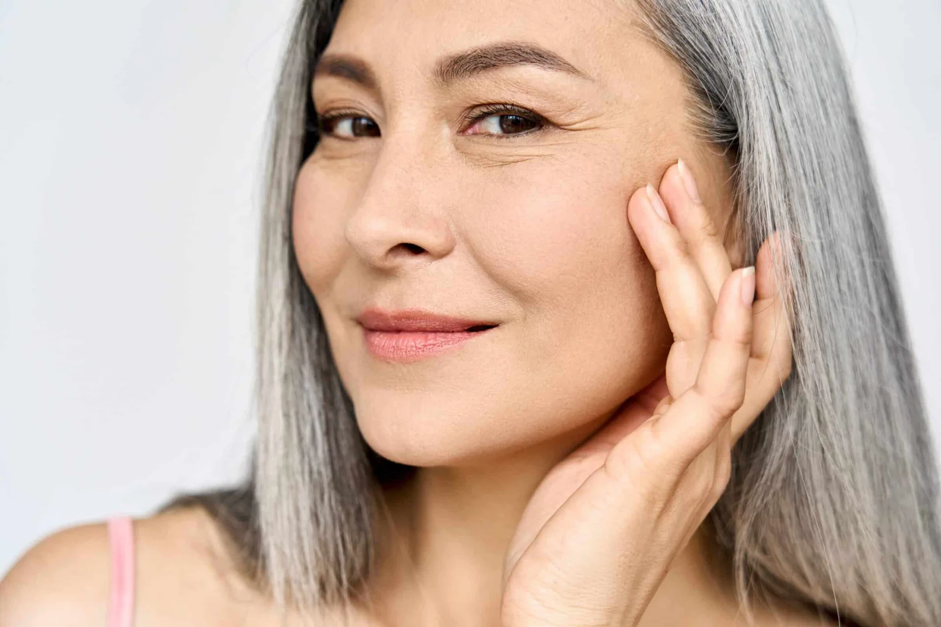 Smiling Asian woman with long gray hair gently touching her cheek, conveying natural beauty