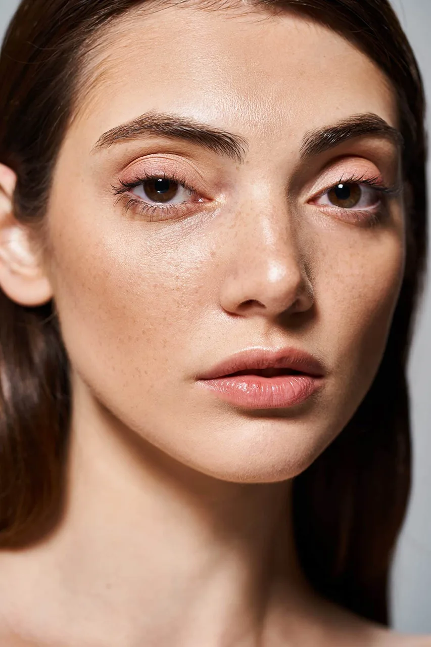 Close-up of a woman's face with brown eyes, natural eyebrows, freckles, and glossy nude lips. She wears subtle pink eyeshadow