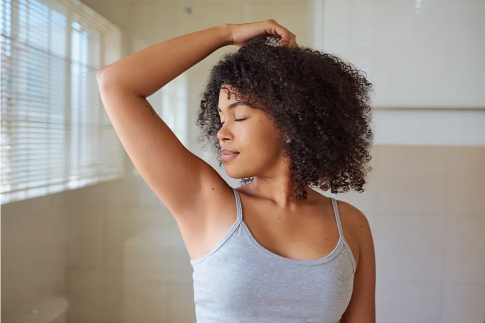 Woman with curly hair and closed eyes lifts her arm, revealing a smooth underarm. She appears fresh and comfortable